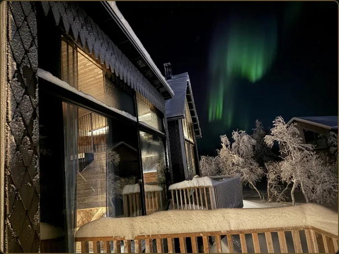 Green aurora streaks over the lit glass windows of an Aura chalet, snow on the deck