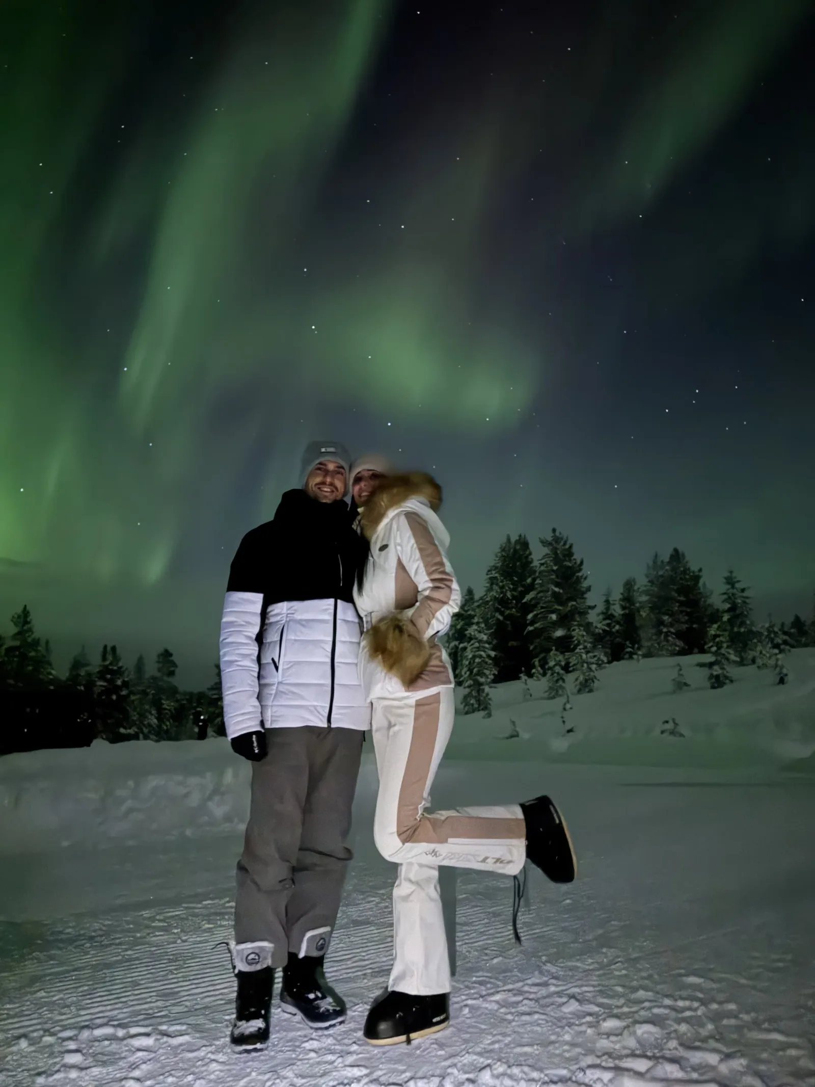 Two guests standing together under the northern lights in the snow at Aura Arctic Chalets