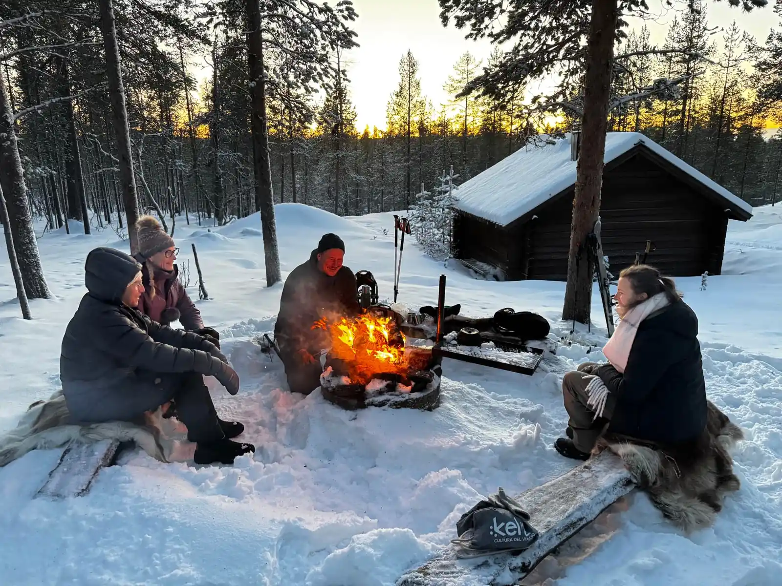Guests gathered around a glowing campfire in a snowy forest at sunset near an Aura chalet